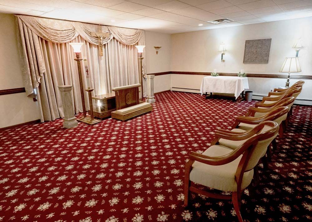 Funeral home interior with red patterned carpet, beige chairs, and drapes near a casket.
