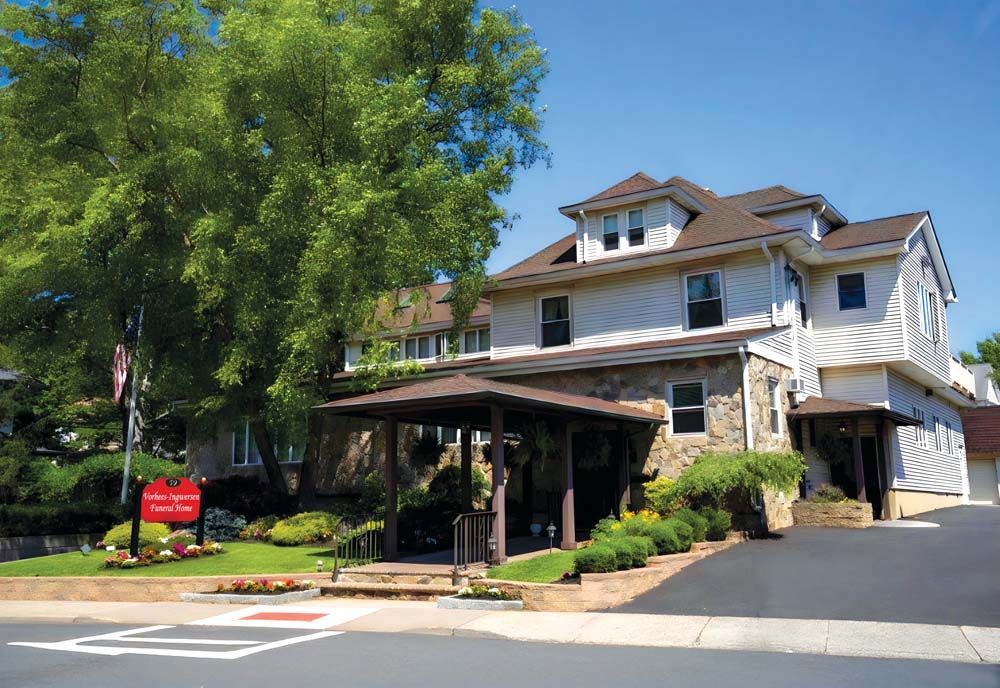 Exterior of a light-colored funeral home with stone accents, under a blue sky with a tree and sign.