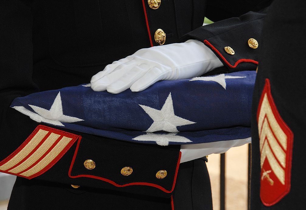 Soldier in uniform holds folded American flag. White-gloved hands, blue, white, and red colors.