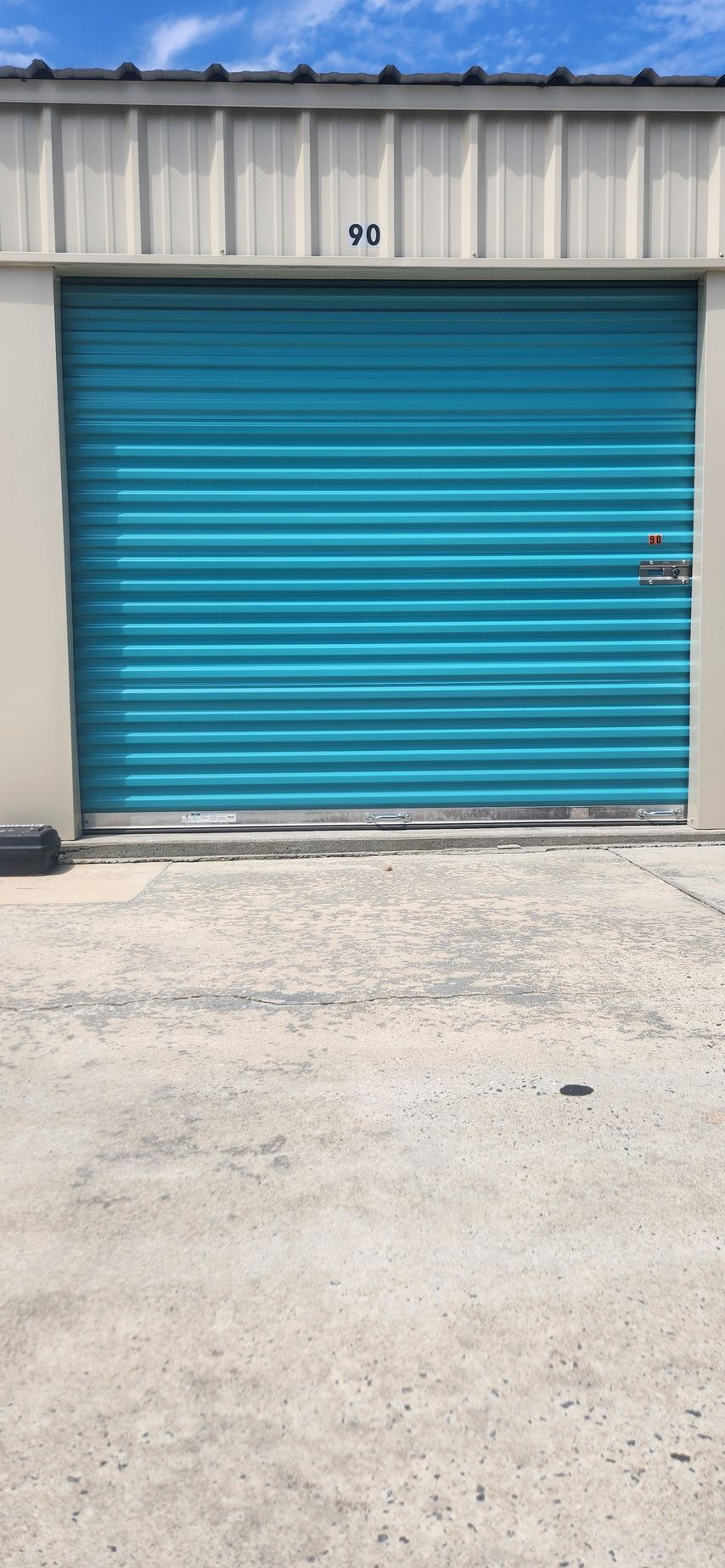 White metal storage units with blue roll-up doors on a gravel lot under a partly cloudy sky