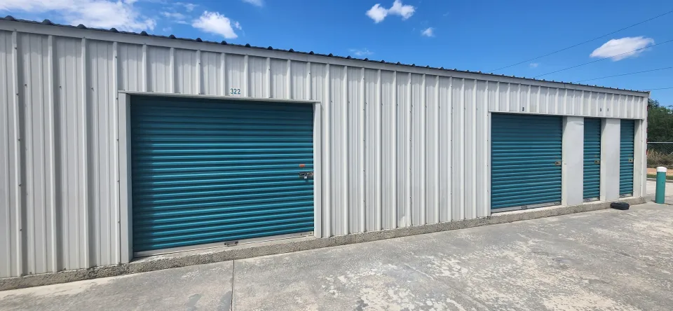 White metal storage units with blue roll-up doors on a gravel lot under a partly cloudy sky