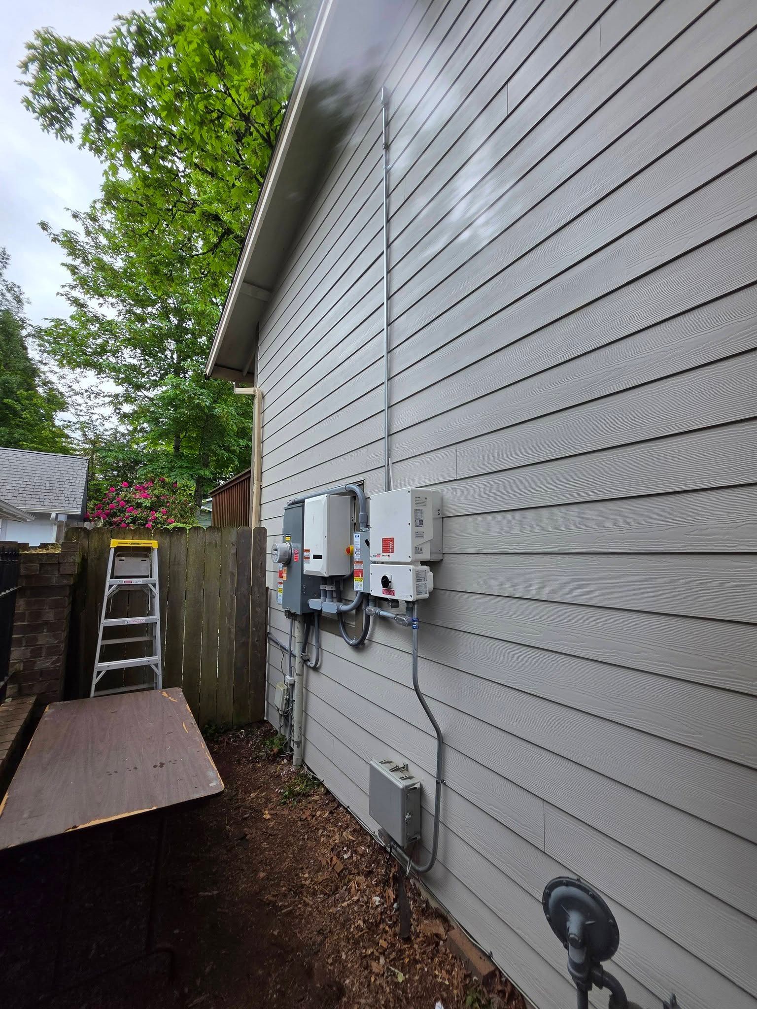 Side yard beside a gray house with utility meters, a fence, ladder, and trees overhead.