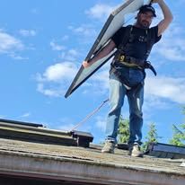 Worker on a roof carrying a solar panel overhead beneath a blue sky