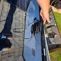Close-up of a blue solar panel on a rooftop, with a hand holding black cables beside it.