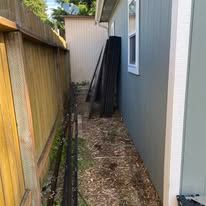 Narrow side yard between a wooden fence and gray house, with stacked black pipes and dry ground cover.