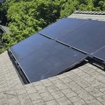Solar panels on a sloped shingle roof beside green trees.