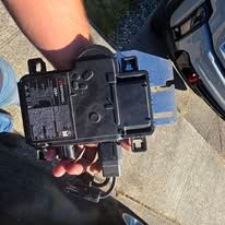 Close-up of a hand holding a black electronic device on a sunlit pavement near a car.