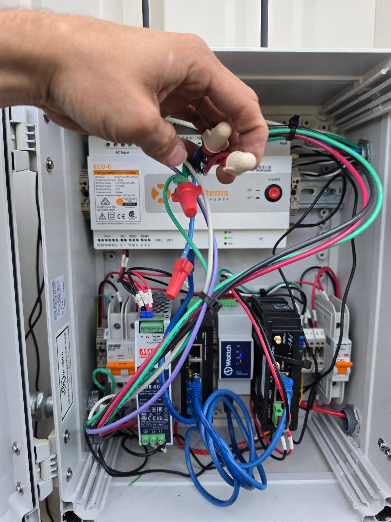 Hand holding wires inside an open electrical control panel with tangled multicolored cables and components