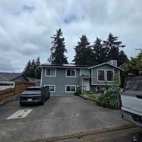 Gray two-story house with driveway, parked cars, and tall trees under a cloudy sky