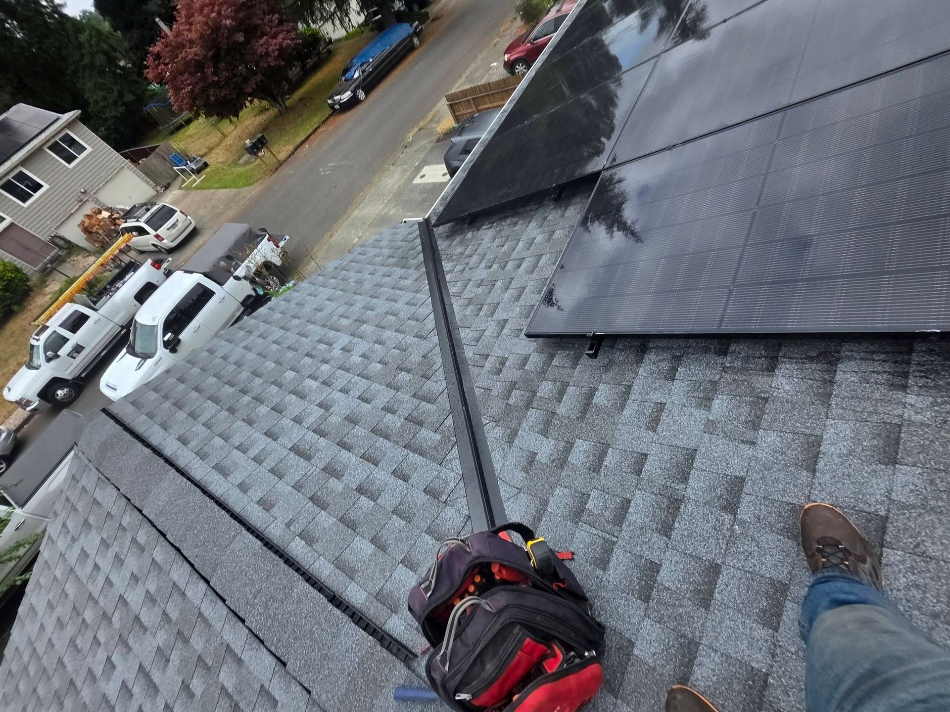Roofing worker on a gray shingle roof with tools, overlooking a residential street.