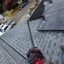 Person on a gray shingled roof using a safety harness, with a ladder and driveway below.