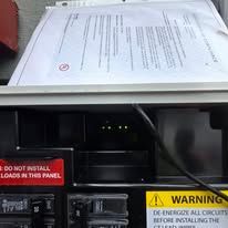 Open electrical panel with warning labels and papers resting on top