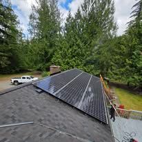 Solar panels installed on a dark shingled roof beside tall green trees and a parked white truck.