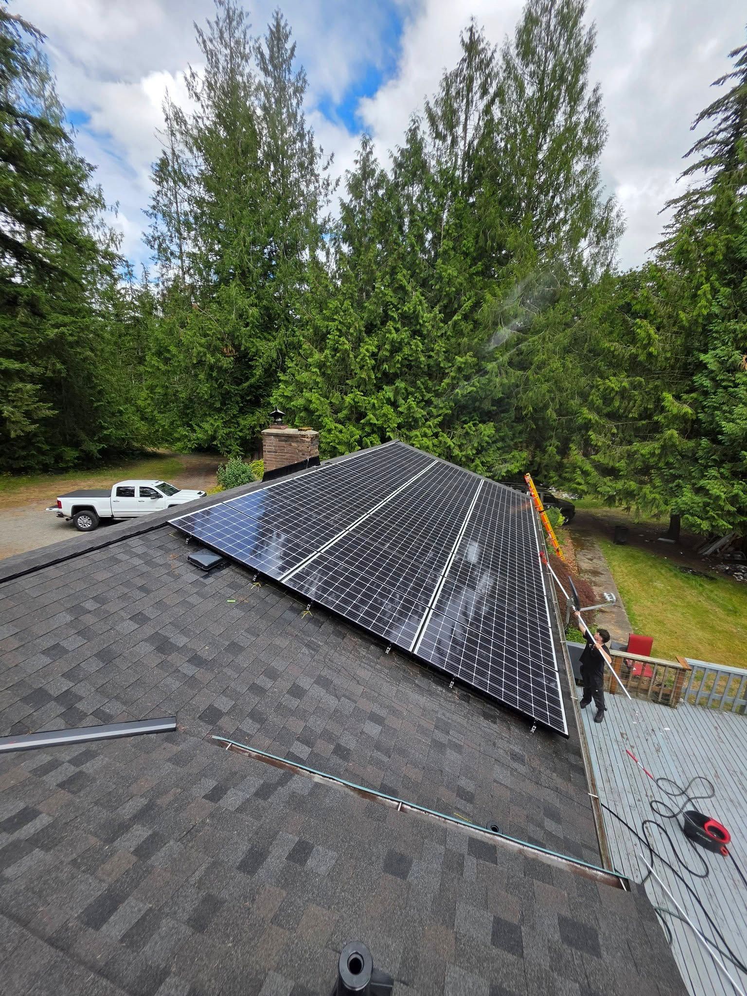Roofers installing black shingles on a steep gray roof, with trees and a truck in the background