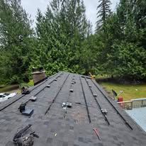 Roof under construction with black shingles and rooftop equipment, backed by tall evergreen trees
