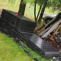 Stacked black solar panels leaning in a grassy yard beside trees and a parked car.