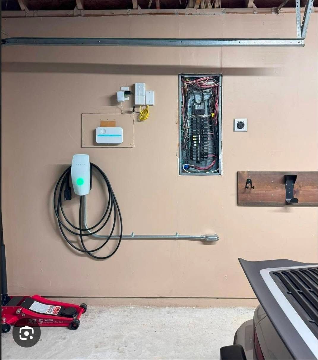 Garage wall with electrical panel, charger cable, outlets, and a mounted pipe.
