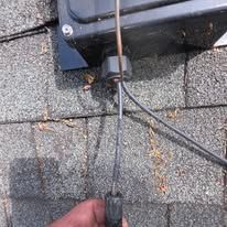 Hand holding a cable at a rooftop roof shingle and vent connection near a black mounting box