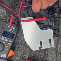 Hand holding red test leads against a metal bracket beside a digital multimeter on gravel