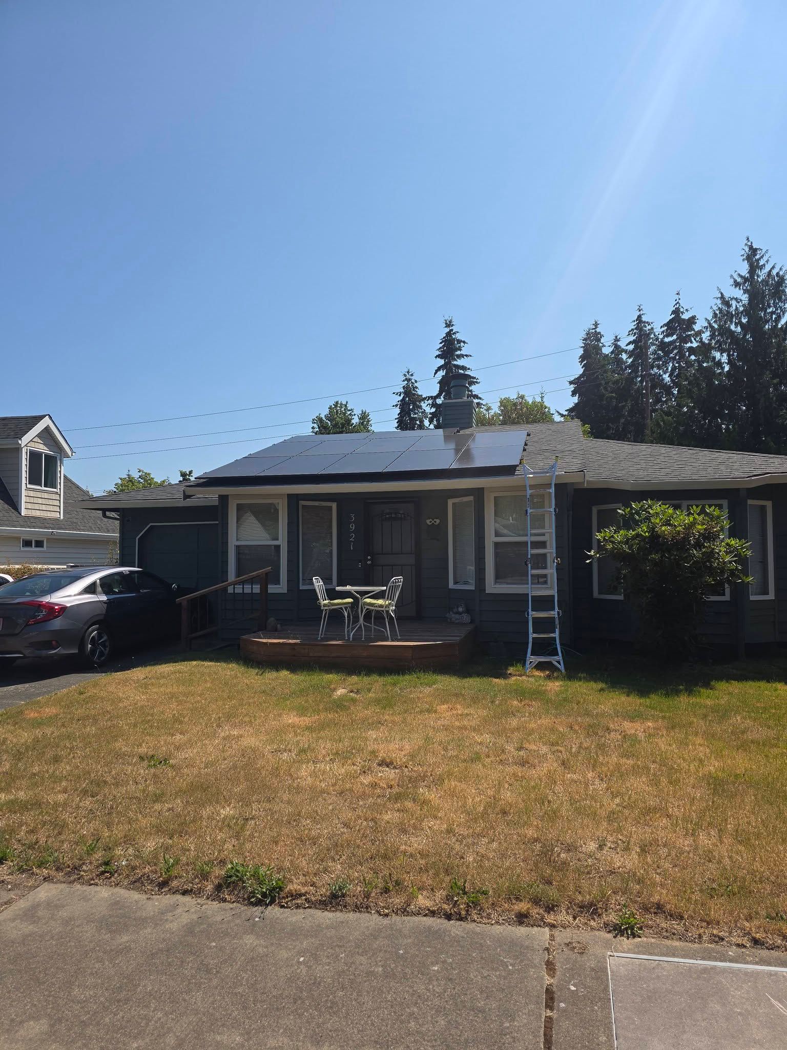Single-story gray house with a small front porch, dry lawn, and parked car under a clear blue sky