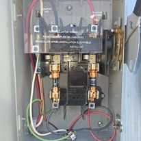 Open electrical panel with wiring, copper pipes, and valves inside a gray metal cabinet