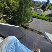 View from a rooftop over gray shingles, with a knee in jeans in the foreground and trees and houses below.
