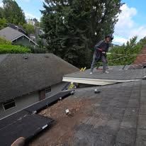 Worker standing on a damaged roof beside a lifted shingle section, with houses and trees in the background