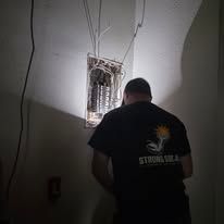 Man in black shirt standing in a dim room, facing an open electrical panel with exposed wires.