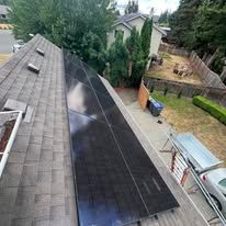 Rooftop solar panels installed on a gray shingled house beside a driveway and fenced backyard.
