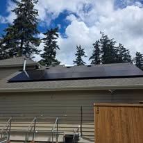 House roof with solar panels beneath a cloudy sky, with tall evergreen trees behind it