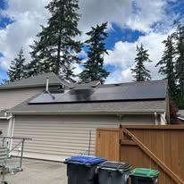 Single-story house with solar panels on the roof beside a wooden fence and trash bins under a cloudy sky