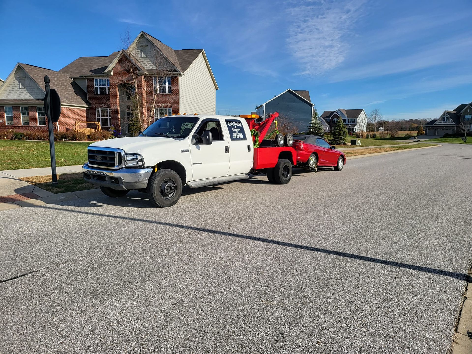 A white tow truck is towing a red car down a street.