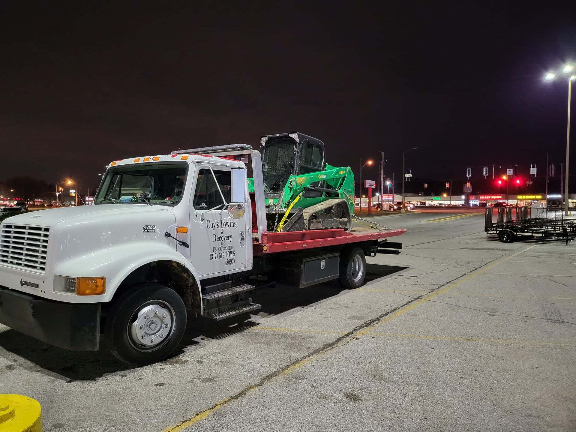 A tow truck with a green excavator on the back is parked in a parking lot at night.