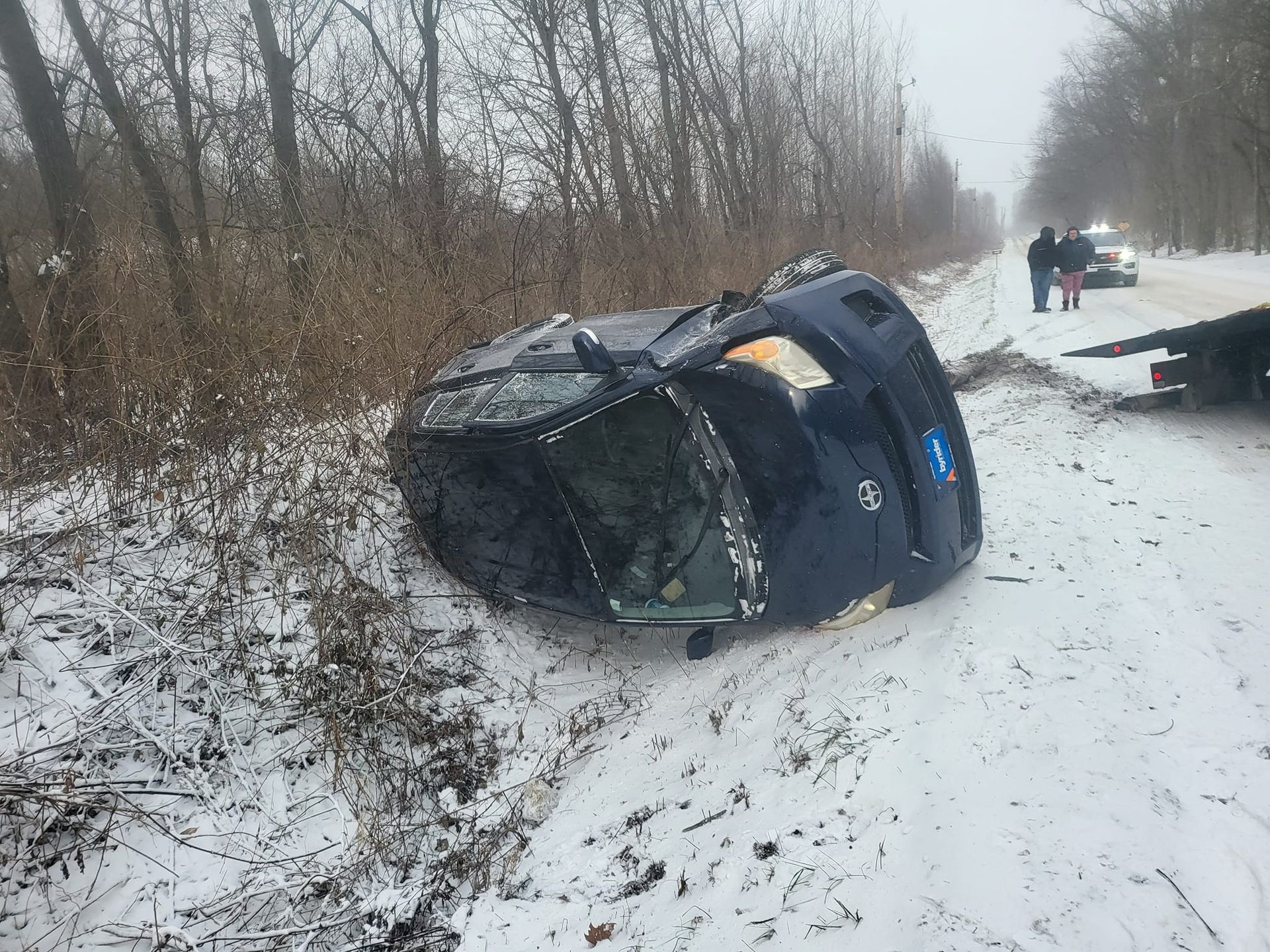 A car is turned over on the side of the road in the snow.