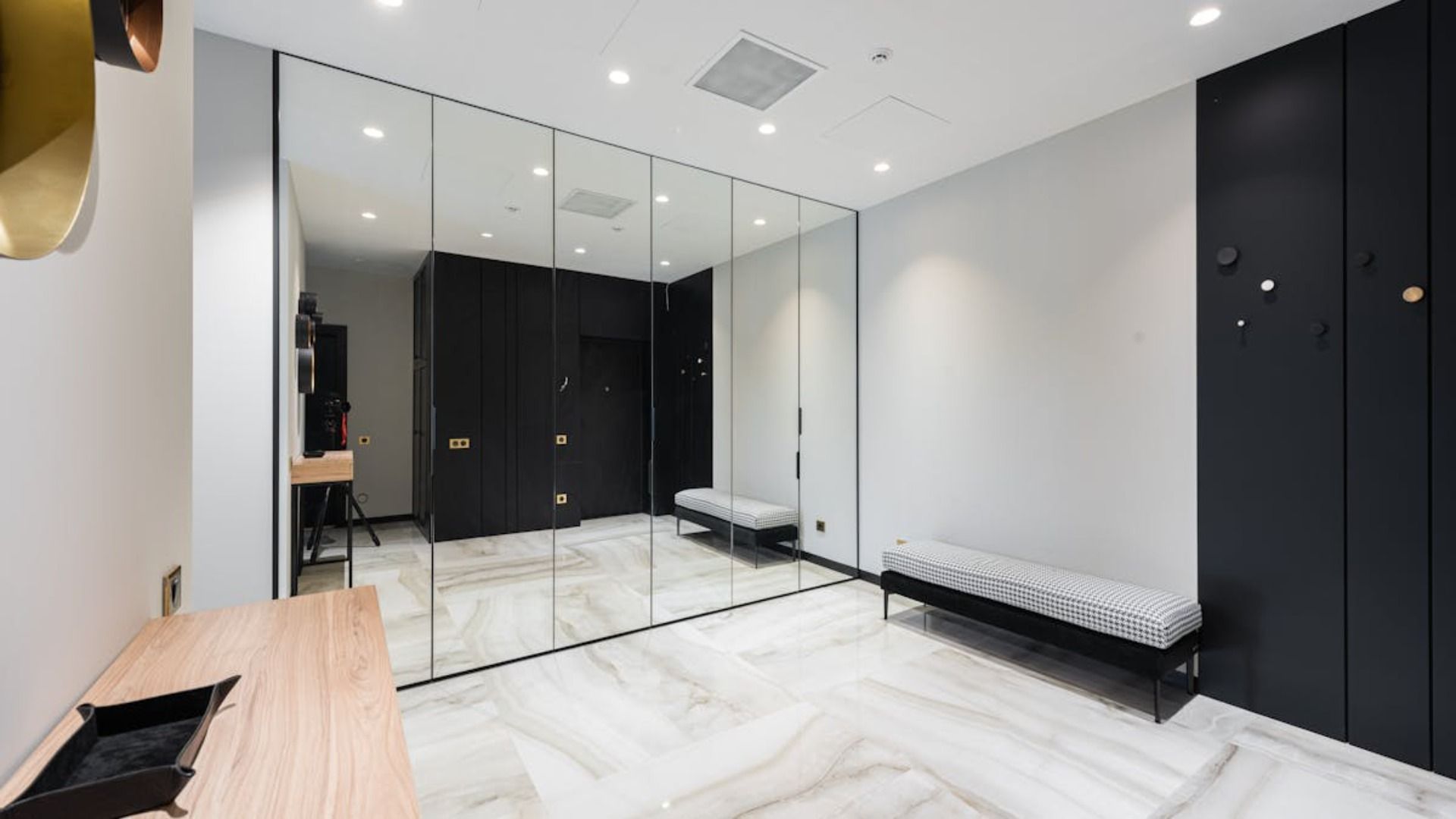 Pasillo minimalista con suelo de mármol claro, un largo banco gris, armarios negros y una pared con espejo de cuerpo entero.