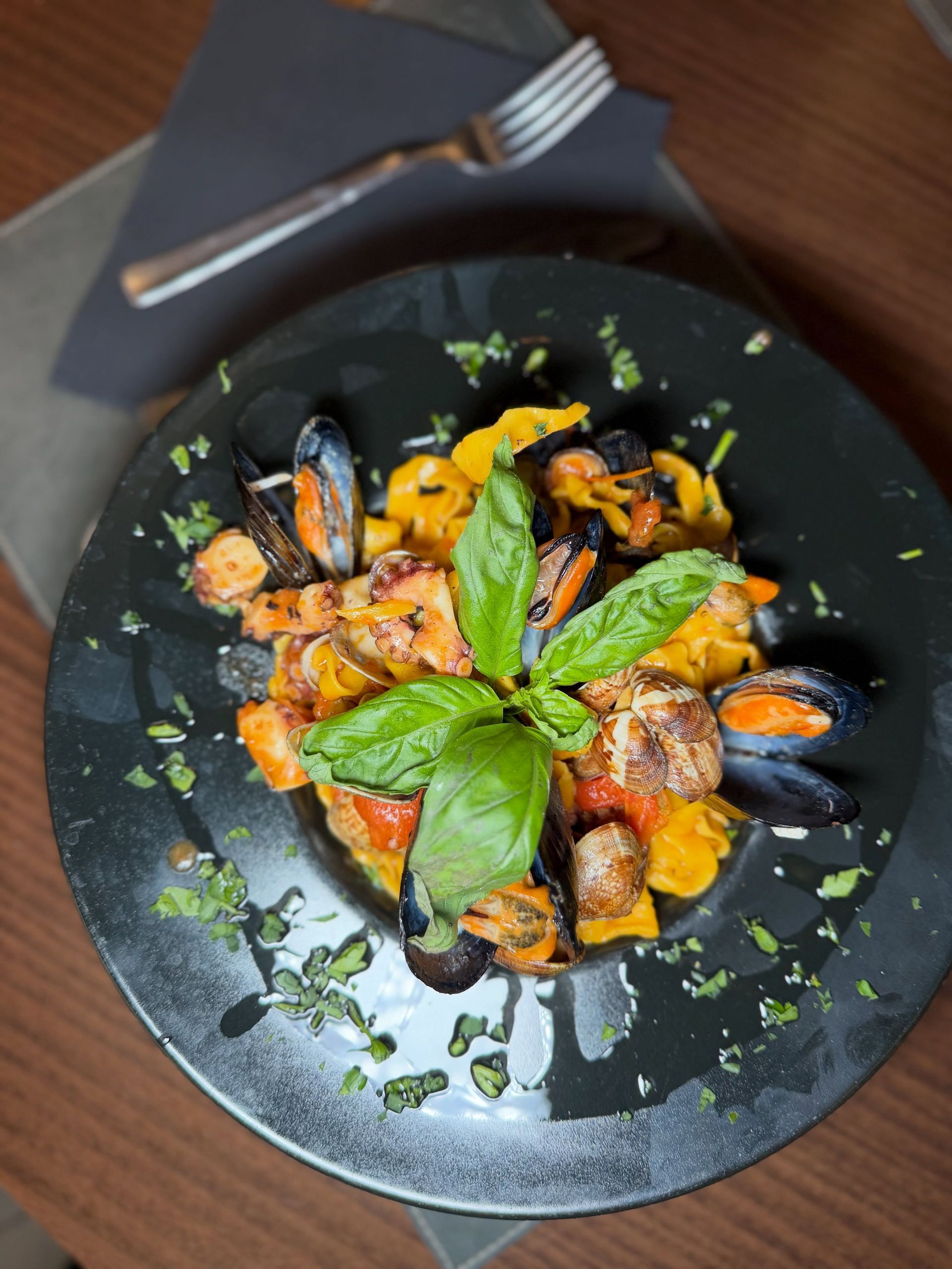 Un piatto nero di cozze e verdure arrostate guarnite con basilico, accanto a una forchetta e un coltello su un tavolo scuro.