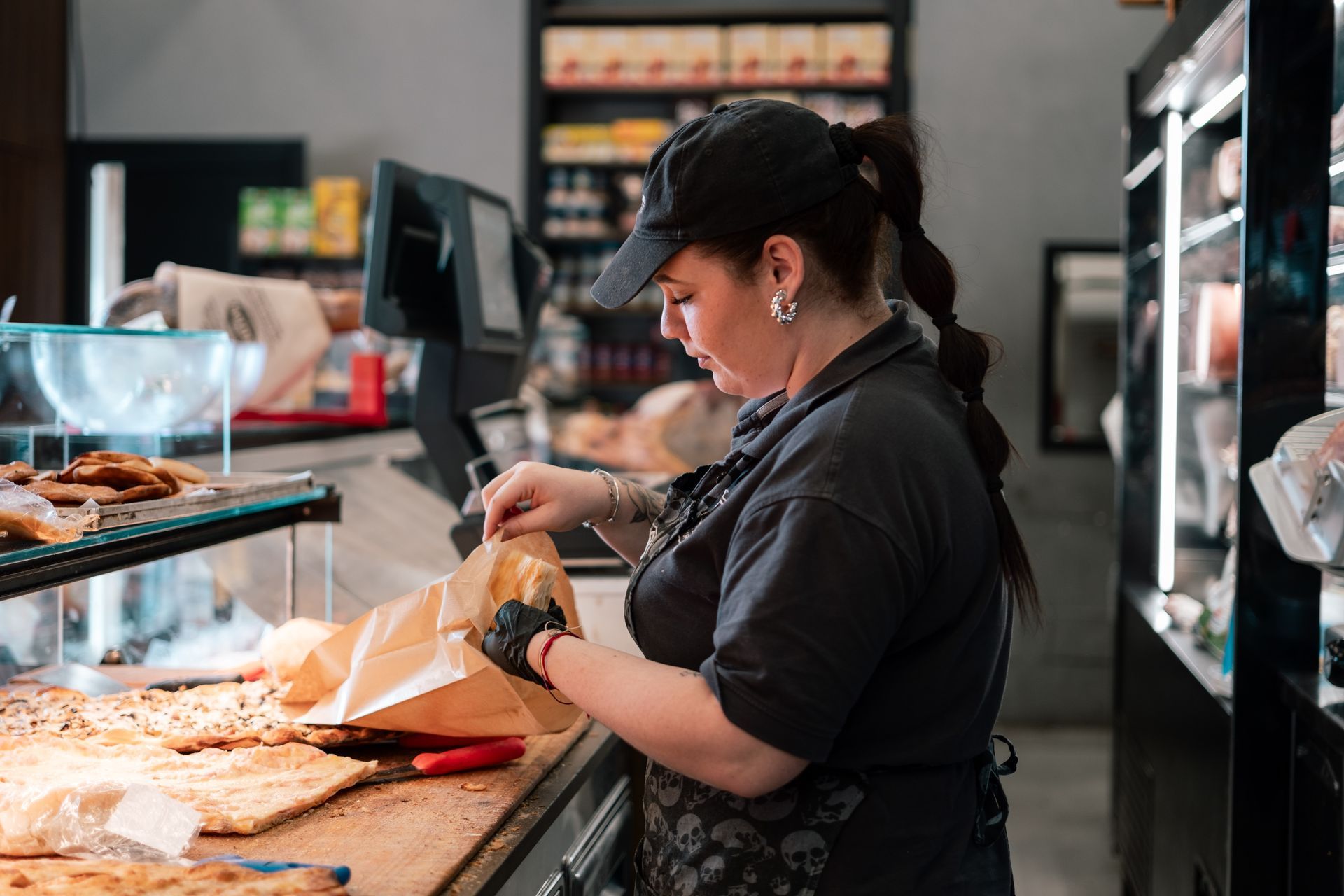 Un'operaia di panetteria confeziona pasticcini al bancone, con vassoi di prodotti da forno e vetrine espositive nelle vicinanze.