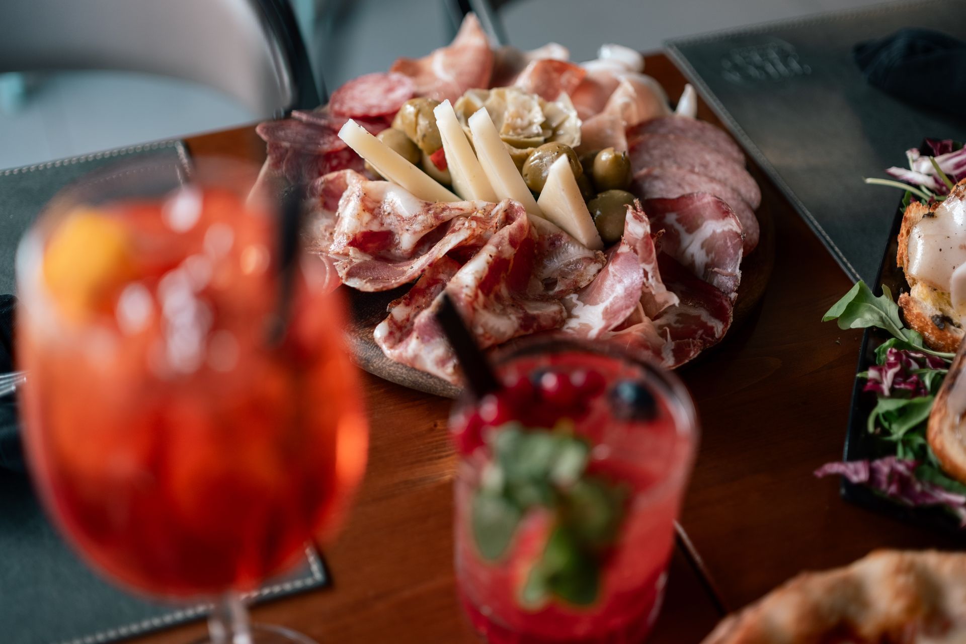 Su un tavolo si trovano assaggi di antipasti e cocktail rossi, con al centro un vassoio di salumi e formaggi.