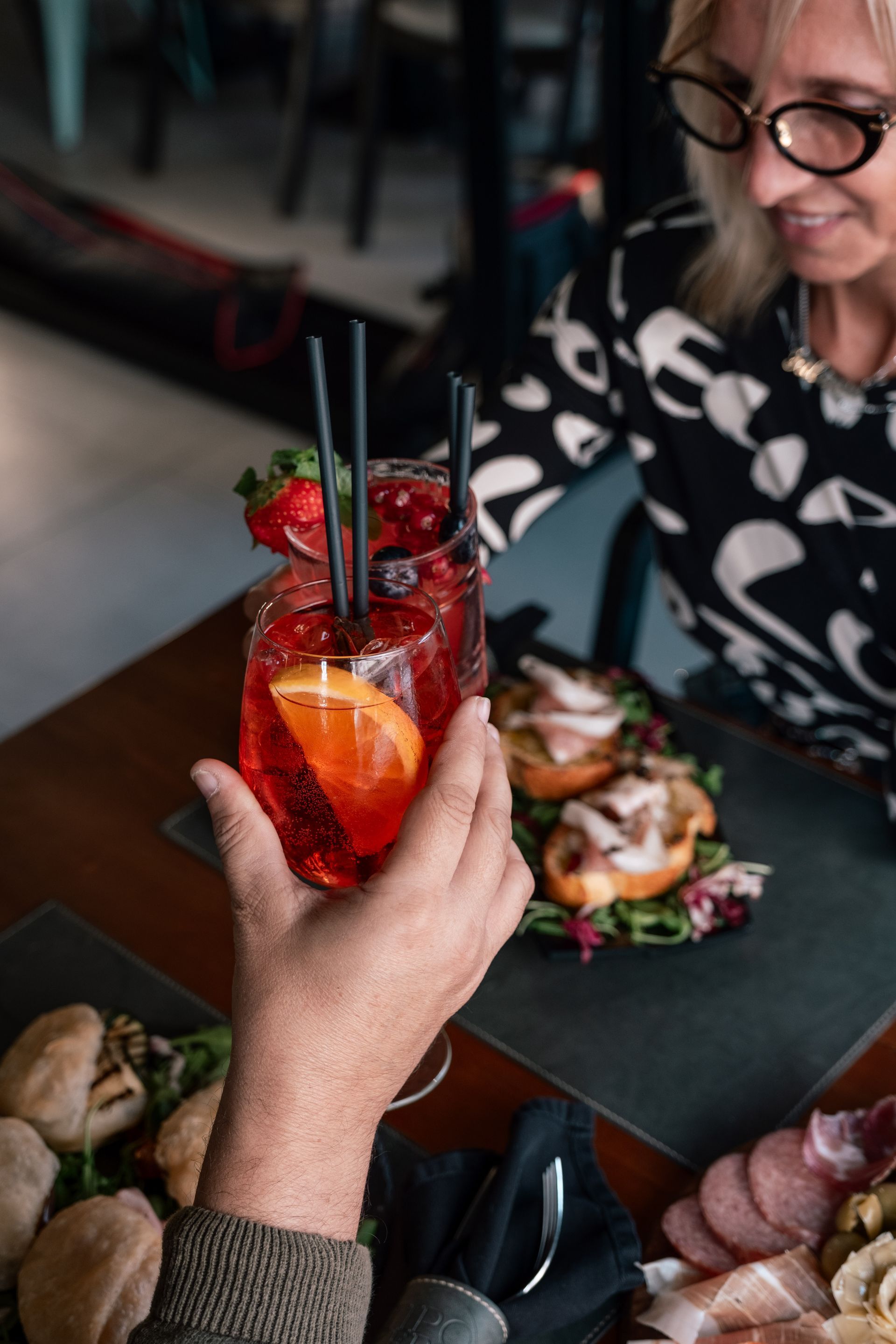 Una mano che regge un cocktail rosso con fragole su un tavolo di ristorante con piatti pronti e un cliente sorridente nelle vicinanze.