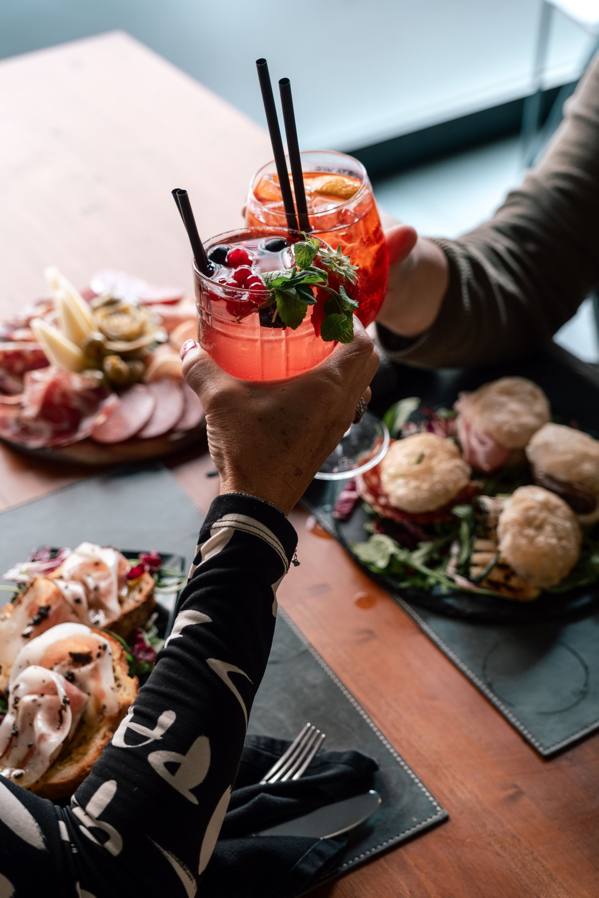 Mani che brindano con cocktail colorati sopra un tavolo con antipasti e piccoli piatti