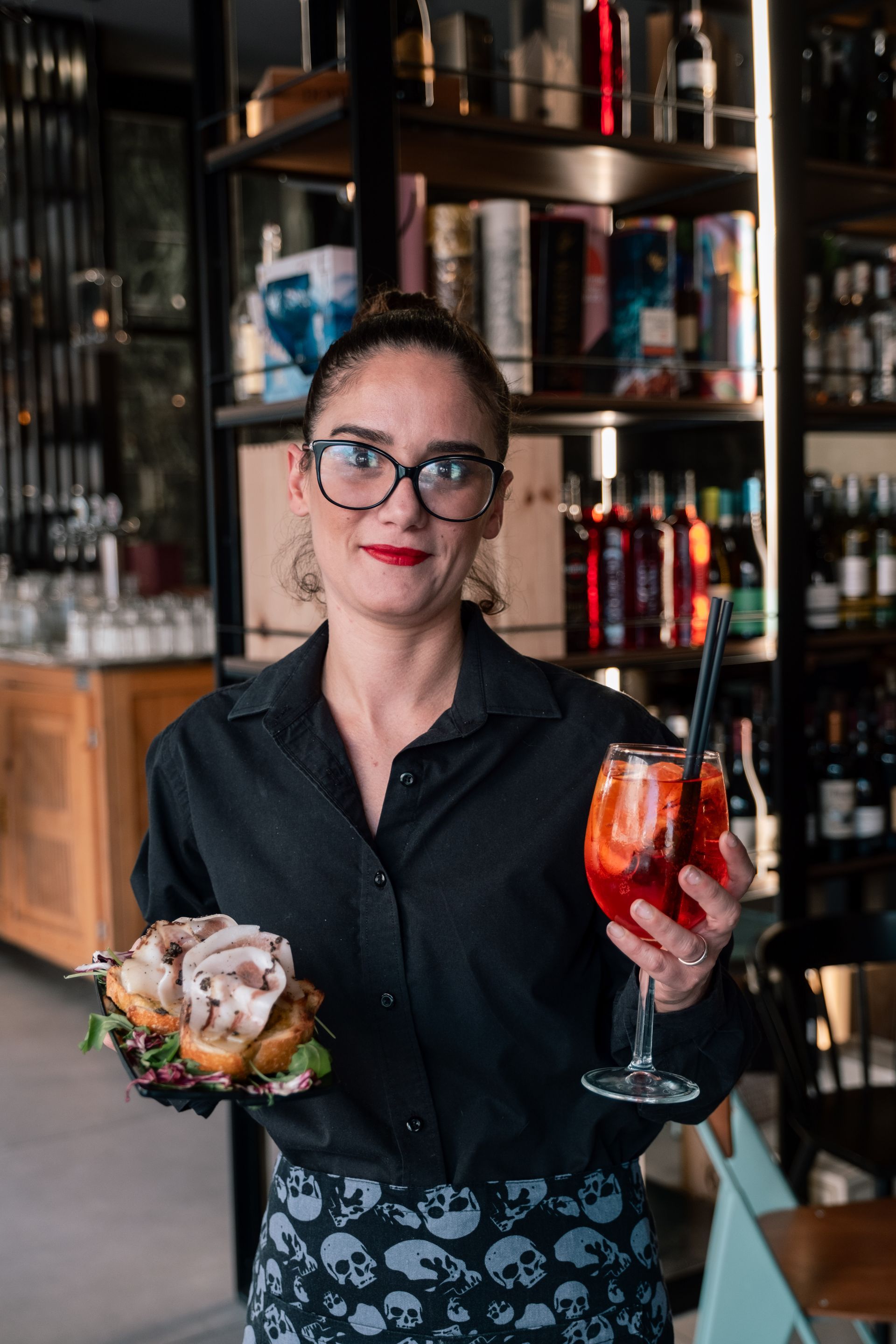 Una donna in un bar tiene in mano un cocktail e un piatto di cibo, in piedi davanti a degli scaffali pieni di bevande.