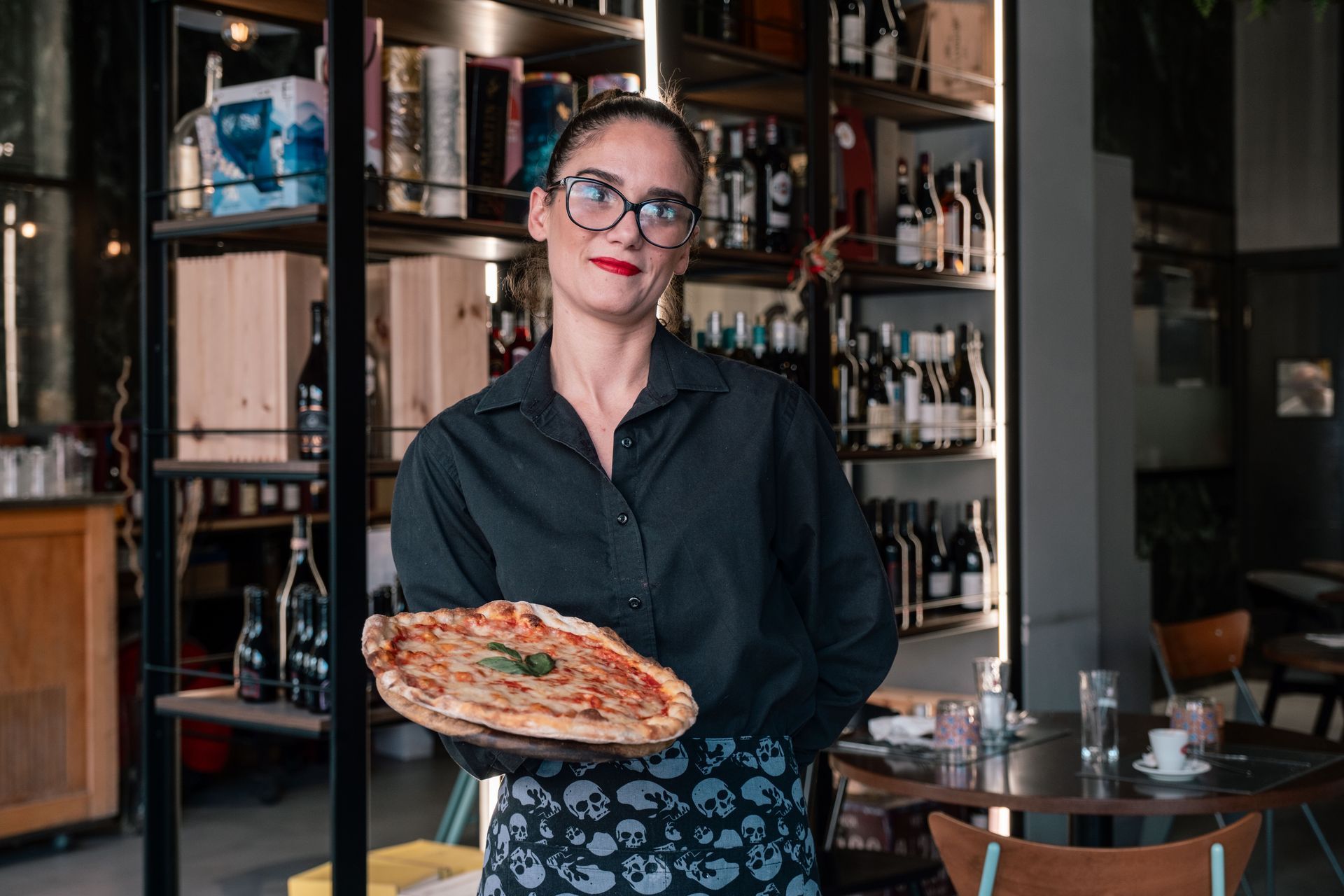 Una donna tiene in mano una pizza in un accogliente ristorante, in piedi davanti a scaffali e tavoli.