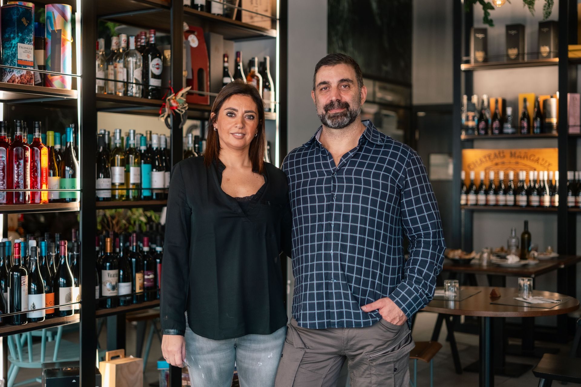 Due persone in piedi in un bar, circondate da bottiglie e bicchieri, sorridono alla macchina fotografica.