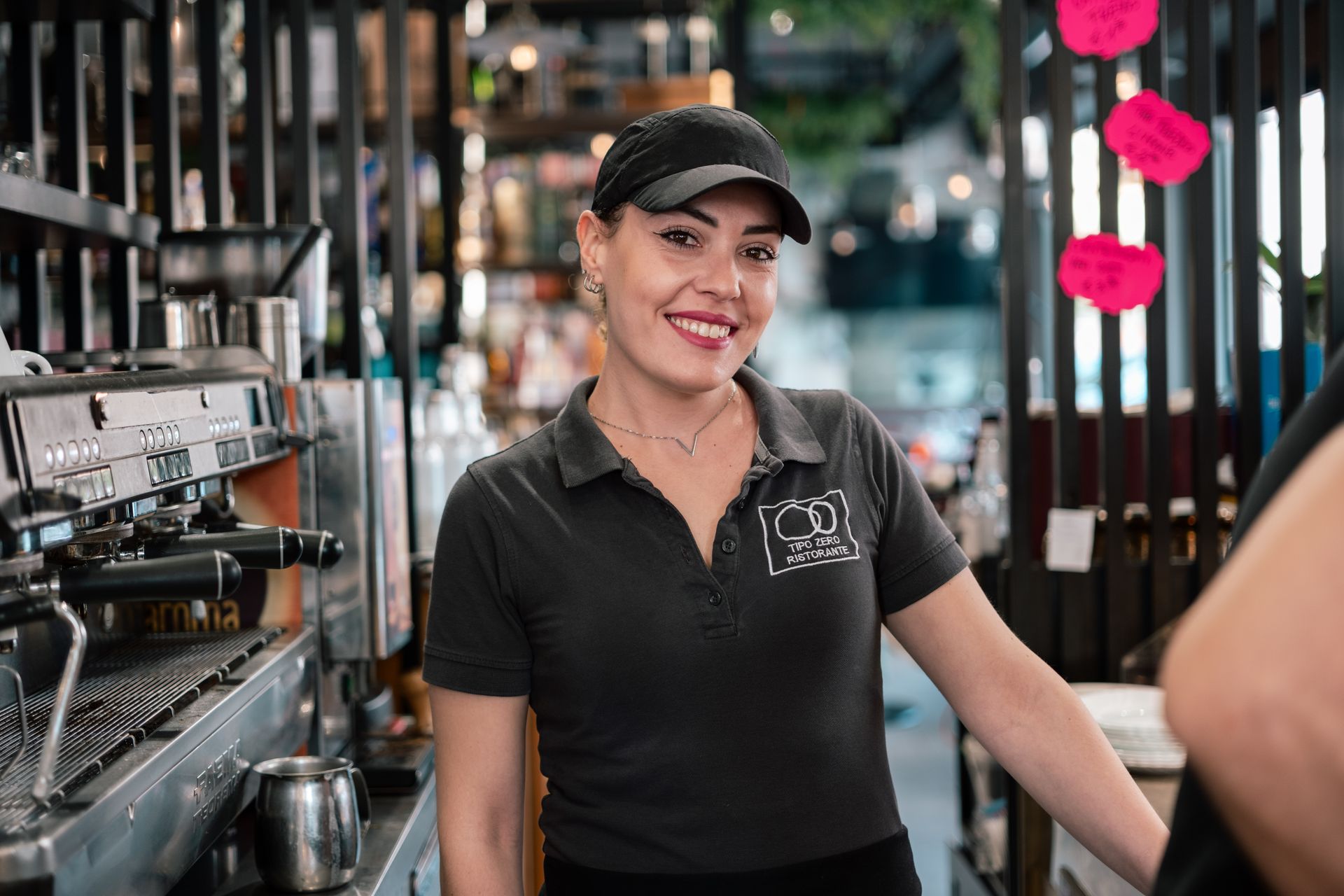 Barista sorridente in uniforme e berretto neri in piedi dietro il bancone del caffè in una caffetteria.