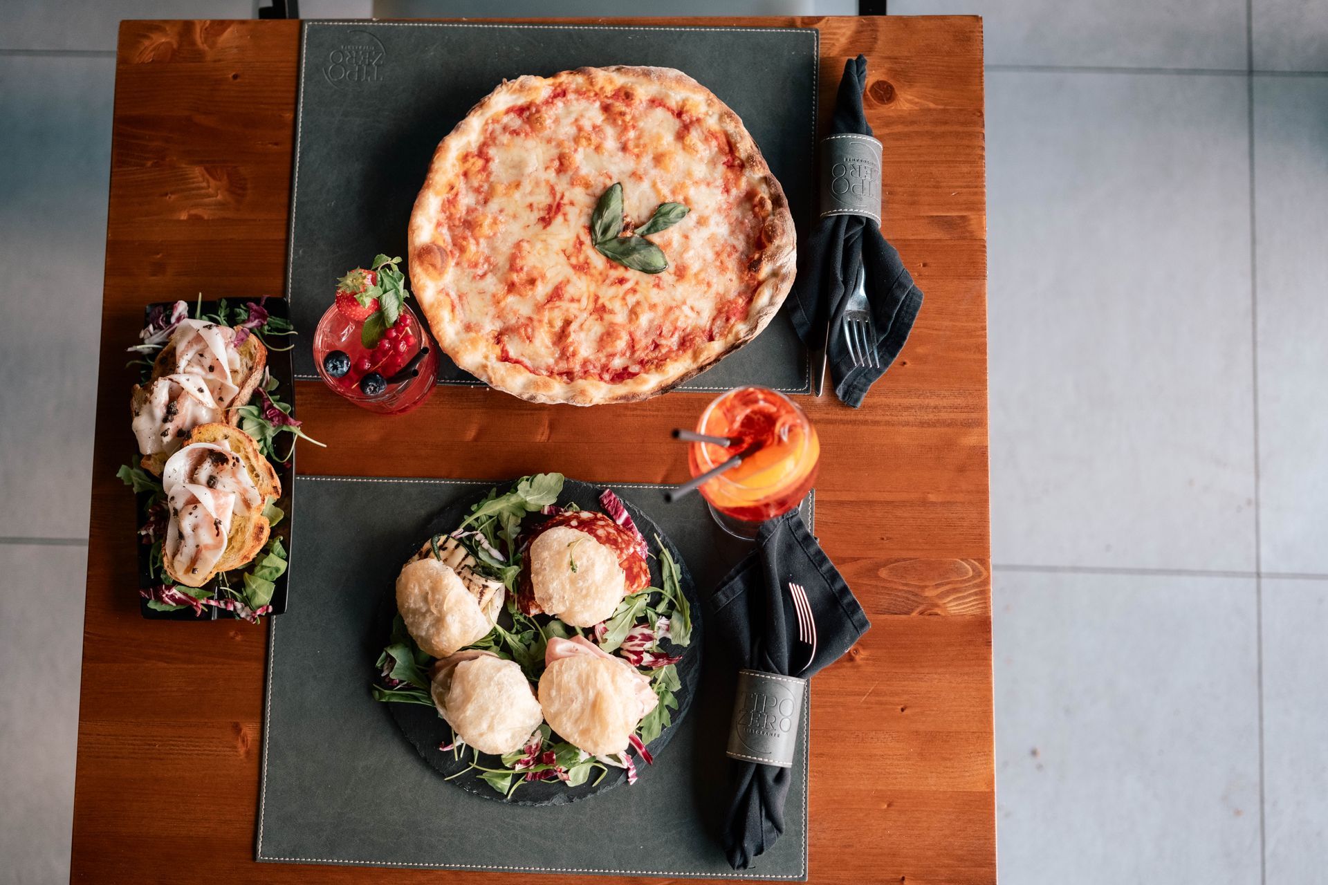 Vista dall'alto di un tavolo di ristorante con pizza, insalata e posate avvolte su tovagliette scure.