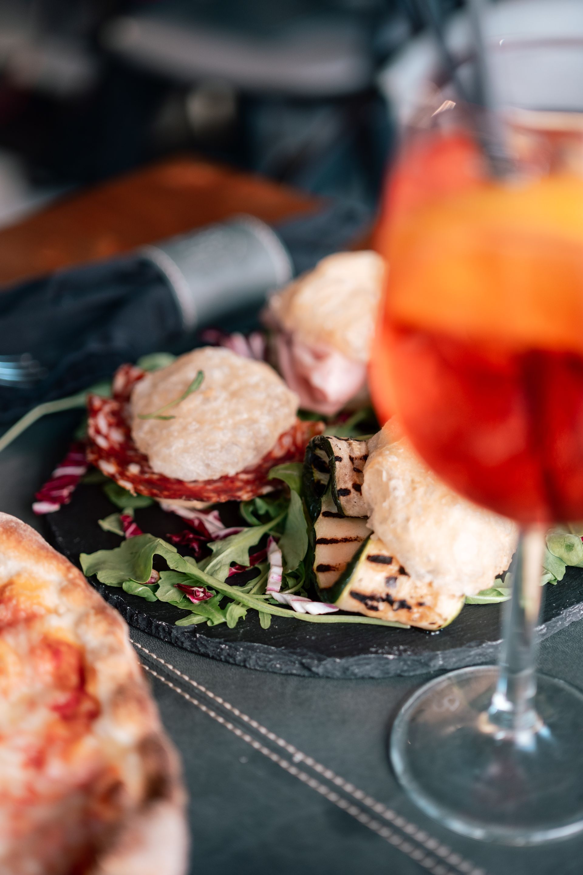 Piatto gourmet impiattato con verdure e pane, accanto a un bicchiere di bevanda rosso-arancio su un tavolo di ristorante.