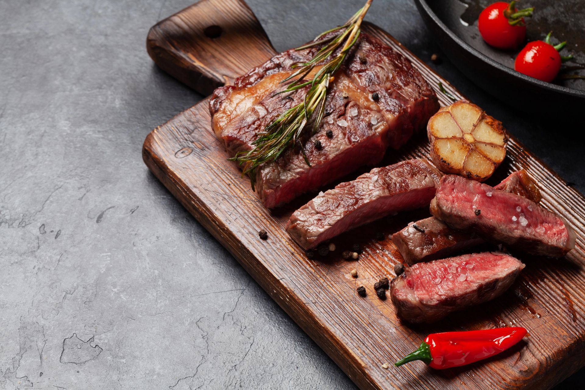 Bistecca alla griglia su un tagliere di legno con rosmarino, carne affettata e pomodorini su un tavolo scuro