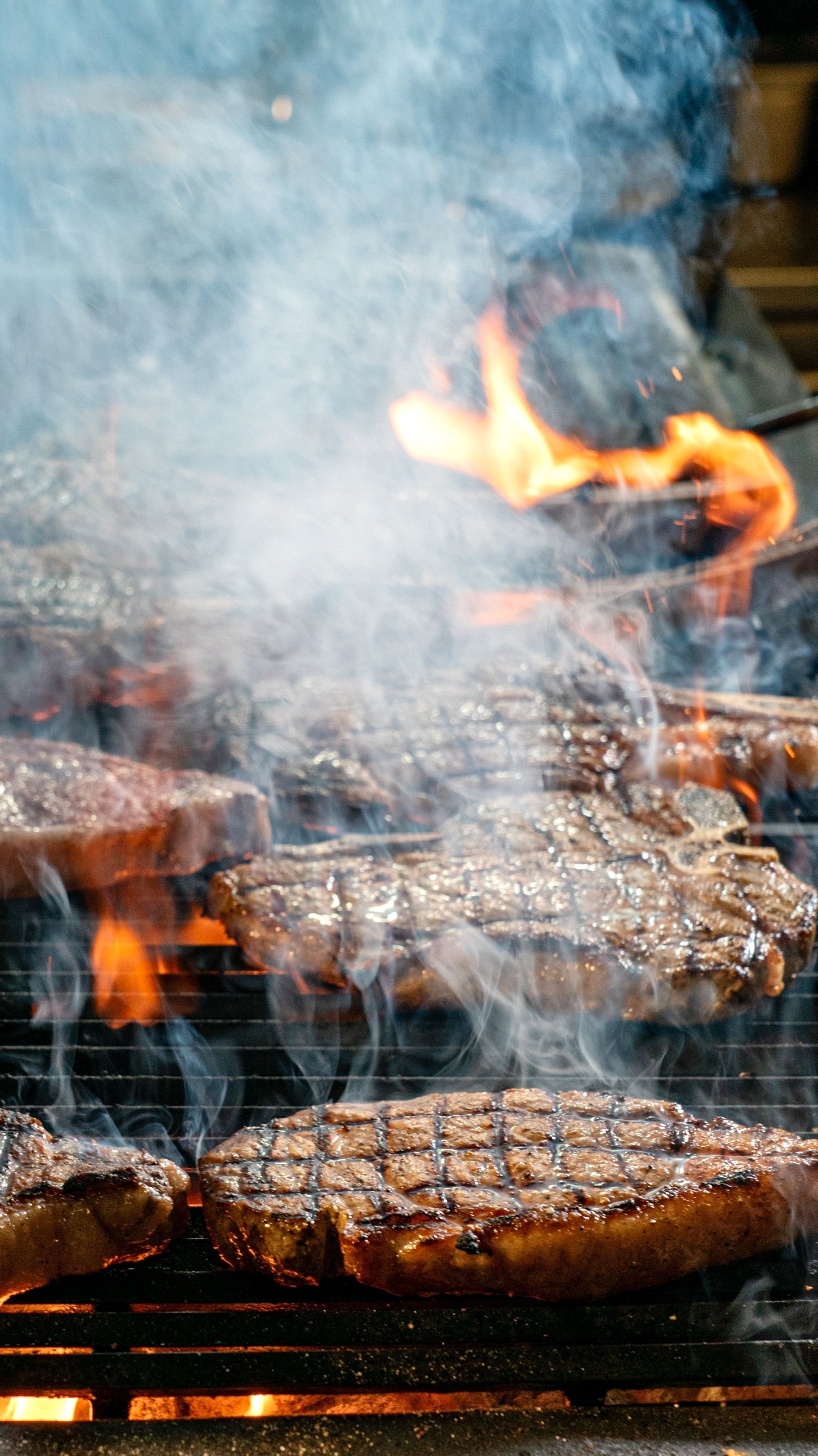 Bistecche grigliate che sfrigolano sulla fiamma viva, con il fumo che sale da una griglia per barbecue.