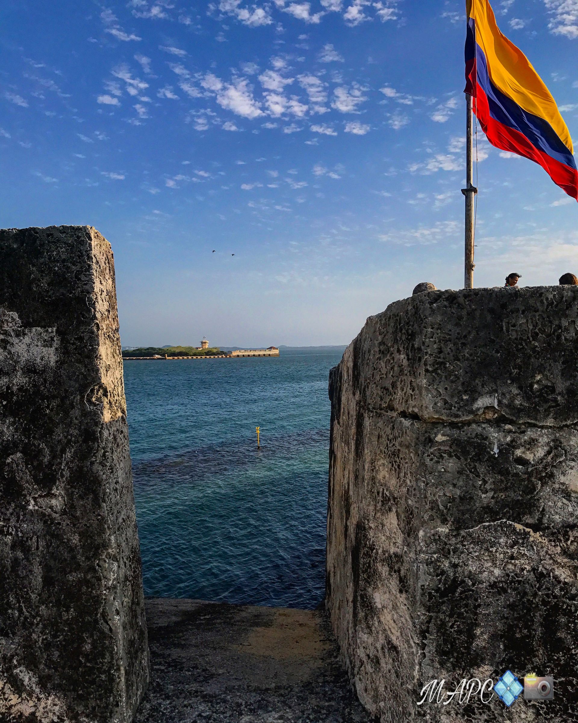 Stone gateway opening to blue sea, with a colorful flag flying on the right under a clear sky.