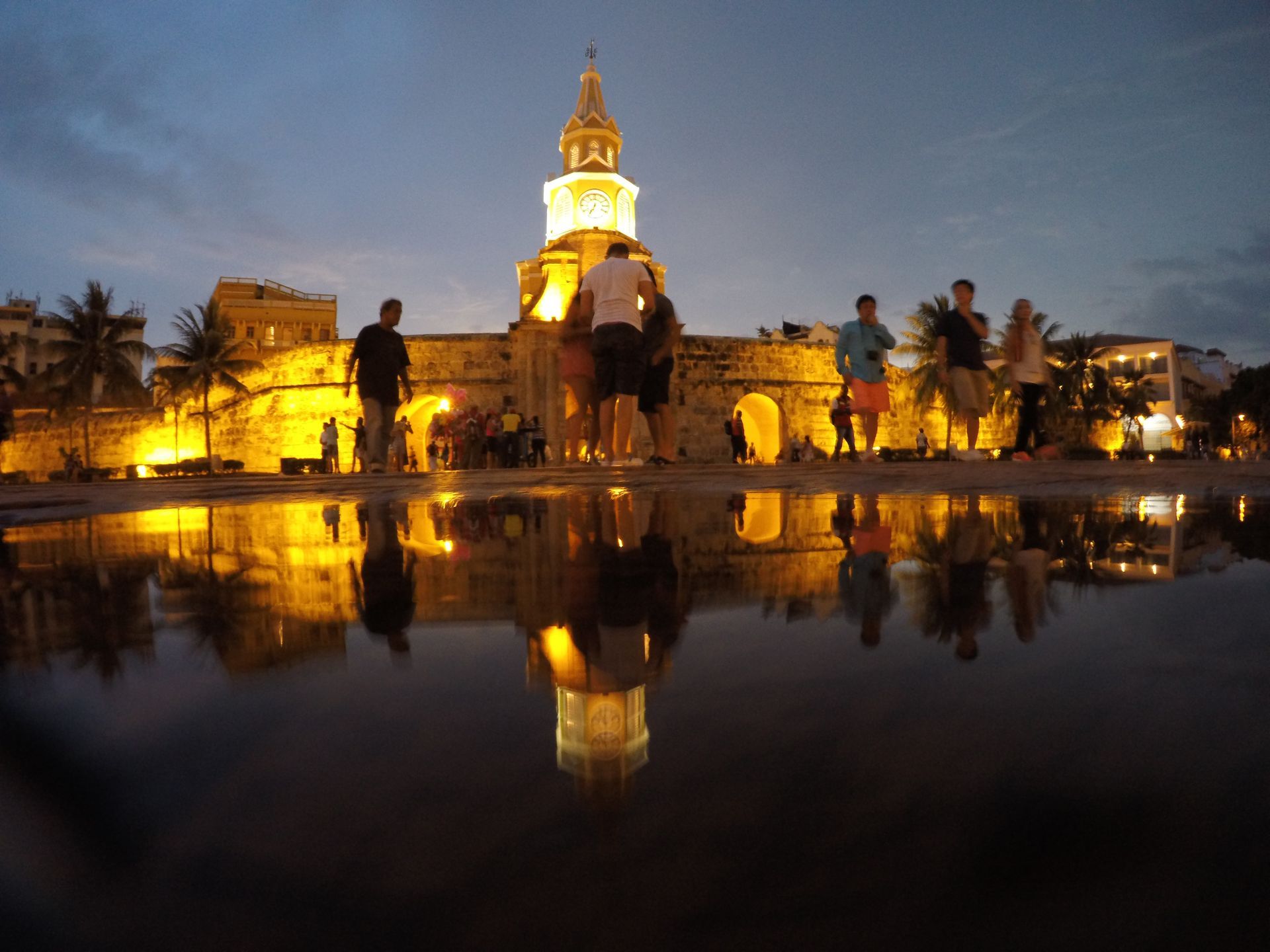 Illuminated church at dusk reflected in still water, with silhouetted trees and people nearby